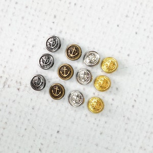 May include: A collection of 12 silver, gold, and bronze anchor buttons. The buttons are arranged in a heart shape on a white background.