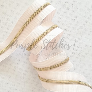 May include: A close-up of a white zipper tape with a gold metal zipper coil. The zipper tape is folded and coiled, showing the detail of the zipper.