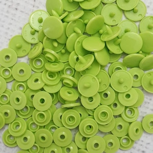 May include: A pile of lime green snap fasteners. The fasteners are made of plastic and have a round, flat top with a protruding post on the underside. The fasteners are used for clothing and craft projects.
