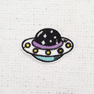 Könnte beinhalten: Ein weißer, blauer und lilafarbener gestickter Aufnäher mit einem Cartoon-UFO mit schwarzem Oberteil und weißen Sternen.