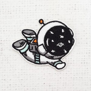 Puede incluir: Parche bordado con un astronauta de dibujos animados en un traje espacial blanco con un casco negro salpicado de estrellas blancas. El parche tiene detalles en gris, naranja y azul claro, y un borde blanco. El astronauta flota en una pose relajada.