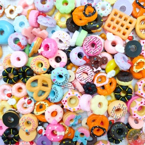 Può includere: Un assortimento colorato di mini ciambelle, pretzel e waffle in vari colori. Le ciambelle presentano glassa rosa, blu e gialla, con zuccherini e scritte al cioccolato. I pretzel sono arancioni e marroni, e i waffle sono marrone chiaro.