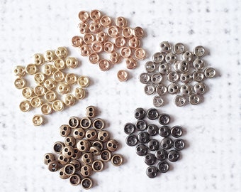May include: A collection of small, two-hole buttons in various metallic finishes, including gold, rose gold, silver, bronze, and black. The buttons are arranged in small clusters against a white background, showcasing their different colors and textures.