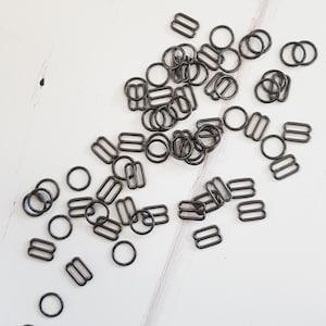 May include: A large quantity of black metal slider buckles, also known as bra strap sliders, are scattered on a white surface. These are commonly used for adjusting the length of straps on clothing and accessories.