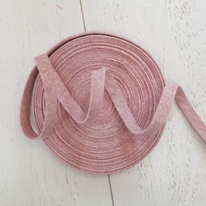 May include: A roll of pink bias binding, with a textured, woven appearance. The binding is coiled in a spiral, with a length extending outwards. The background is a light-coloured wooden surface.