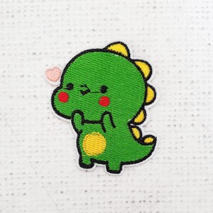 May include: An embroidered patch featuring a cartoon dinosaur in green with a yellow belly, red cheeks, and a pink heart above its head.