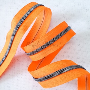 May include: A close-up of a bright orange zipper with a silver metal coil. The zipper is coiled and ready to be used for sewing projects.