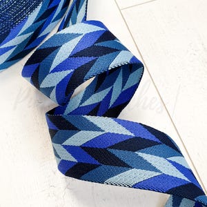 Può includere: Un nastro blu a fantasia con un motivo geometrico. Il nastro presenta un motivo ripetuto di forme a diamante in varie tonalità di blu, tra cui blu navy, reale e azzurro. Il nastro è arricciato e giace su una superficie bianca.