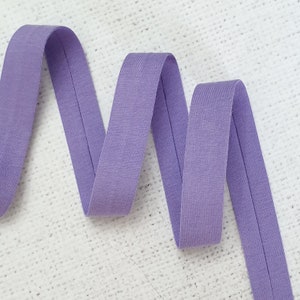 Puede incluir: Cinta elástica lavanda doblada en zigzag. La cinta tiene una textura sutil y una costura central. El fondo es una superficie blanca texturizada.