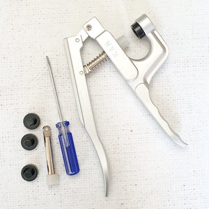 May include: A silver metal tool with a spring and a handle. The tool is used for setting eyelets and snaps. There are three black eyelets, a silver tool, and a blue screwdriver.