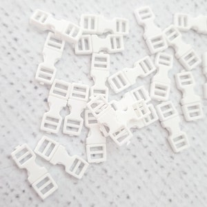 May include: A pile of white plastic slider buckles. The buckles are small and have two holes on each side.