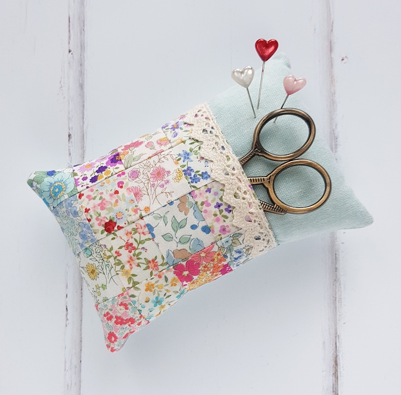 May include: A light blue pincushion with a floral patchwork design and a lace trim. It has a pair of gold scissors and three heart-shaped pins on top.