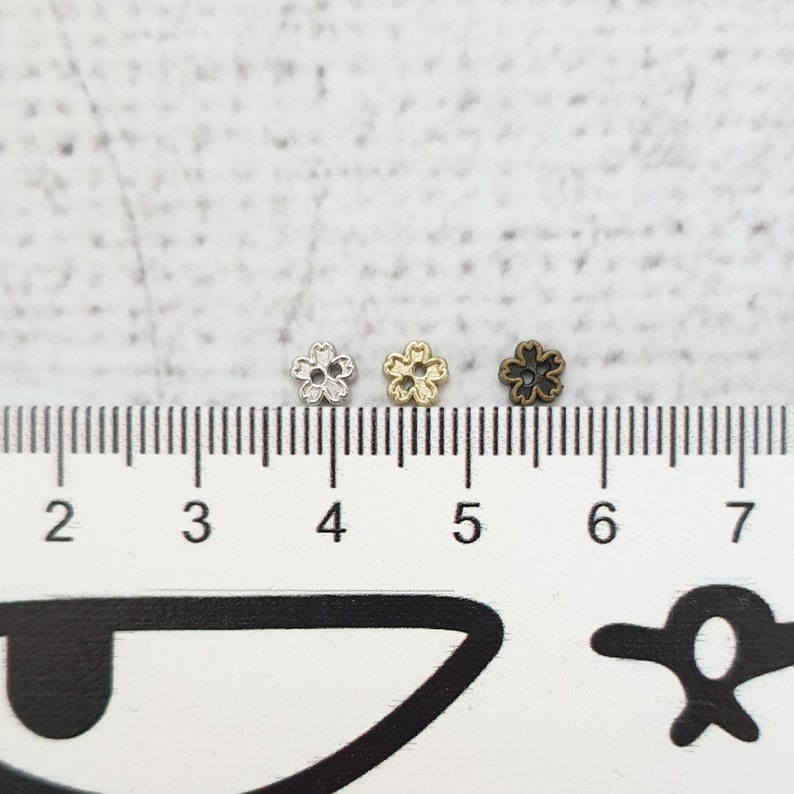 May include: Three small metal flower-shaped charms in silver, gold, and black, measured against a ruler.