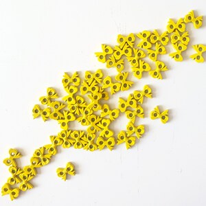 May include: A collection of small, bright yellow wooden bow tie embellishments scattered on a white surface. The bows have a simple, symmetrical design with a small hole in the center of each. The bows are likely for crafting or decorative purposes.