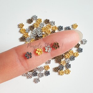May include: Assortment of small metal flower charms in gold, silver, and bronze colors. The charms have five petals and a center with two small red dots. The charms are scattered on a white surface and a finger is holding three charms.