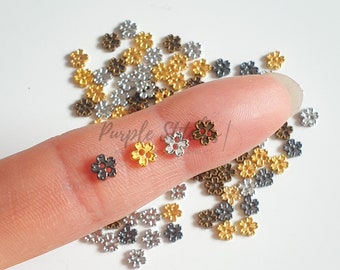 Könnte beinhalten: Sortiment kleiner Blumen-Anhänger aus Metall in Gold, Silber und Bronze. Die Anhänger haben fünf Blütenblätter und eine Mitte mit zwei kleinen roten Punkten. Die Anhänger sind auf einer weißen Oberfläche verstreut und ein Finger hält drei Anhänger.