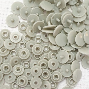 Puede incluir: Una gran cantidad de botones a presión de plástico gris claro. Los botones tienen una forma plana y redonda con un pequeño agujero central y un poste central elevado.