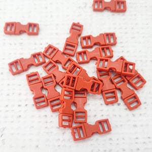 Puede incluir: Un grupo de 18 hebillas deslizantes de plástico naranja. Las hebillas son todas del mismo tamaño y forma, y están dispuestas en un patrón aleatorio.