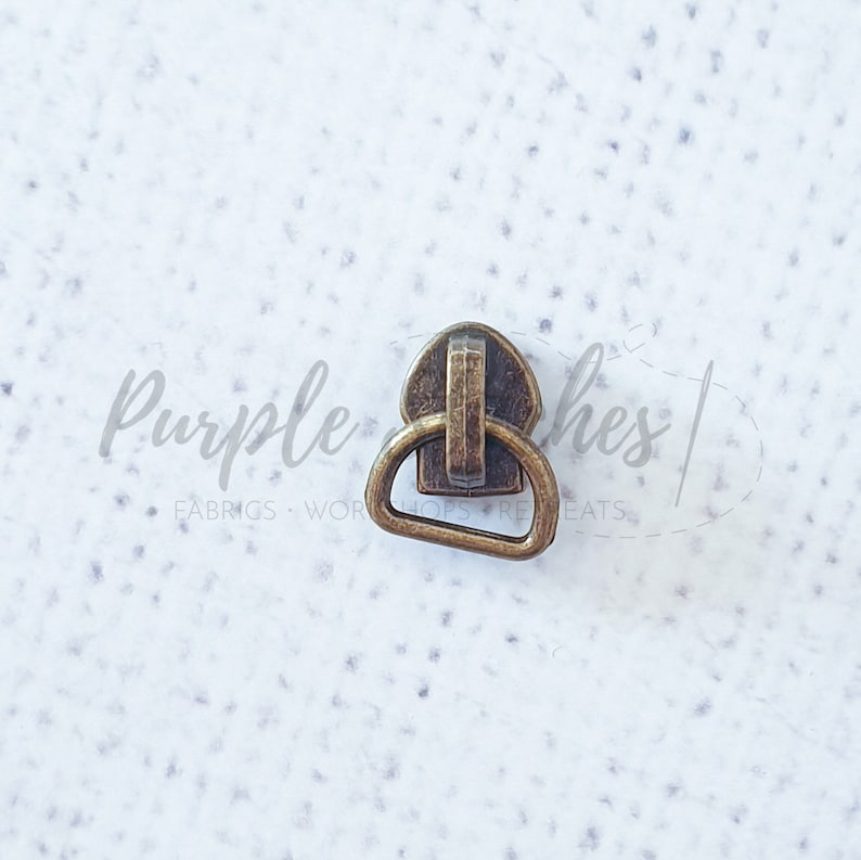 May include: A gold-toned metal zipper pull with a D-ring. The zipper pull is designed to be used with a zipper on a garment or bag.
