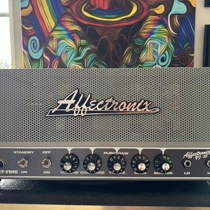 May include: A silver guitar amplifier with the name "Affectronix" printed on the front. The amplifier has a variety of knobs and switches, including a "Kerry-Fine" switch, a "Standby" switch, a "Volume" knob, a "Compress" knob, a "Treble" knob, a "Bass" knob, a "Master" knob, and a "Push Raw" switch. The amplifier also has two output jacks labeled "Lo" and "Hi".