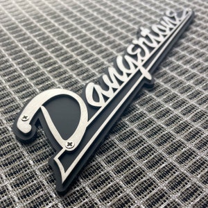 May include: A silver metal "Danelectro" logo with a black background. The logo is attached to a surface with screws.