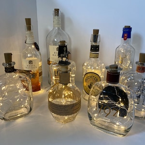 Puede incluir: Una colección de botellas de vidrio con luces de cadena en su interior. Las botellas son transparentes y tienen varias etiquetas. Algunas de las botellas tienen tapones de botella decorativos. Las botellas están dispuestas sobre una superficie blanca.