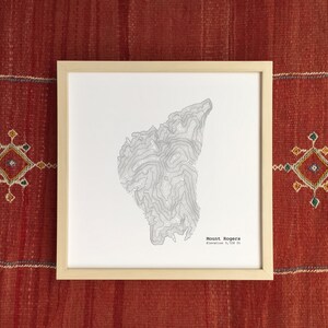 Può includere: Una stampa incorniciata del Monte Rogers, con una mappa topografica dettagliata in tonalità di grigio. La stampa è in una cornice di legno chiaro e include il nome della montagna e l'altitudine, 1.746 m. L'opera d'arte è appoggiata su un tessuto rosso tessuto.