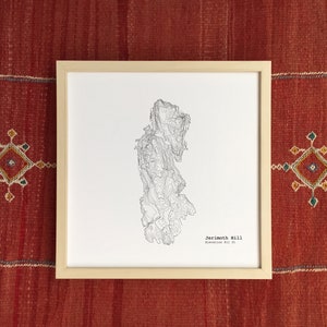 Könnte beinhalten: Gerahmter schwarz-weißer topografischer Kartendruck von Jerimoth Hill, Höhe 247 m. Die Karte befindet sich in einem hellen Holzrahmen, vor einem roten gewebten Hintergrund mit geometrischen Mustern.