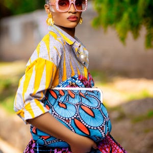 May include: A woman wearing a yellow and blue striped shirt with a large pearl necklace and a blue and orange patterned clutch bag. She is wearing pink sunglasses with rhinestones.