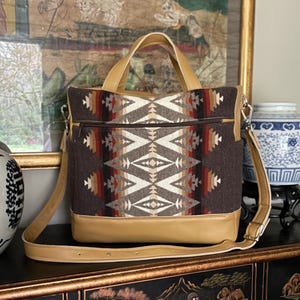 May include: A brown and tan tote bag with a geometric pattern, featuring a tan leather base, handles, and shoulder strap. The bag has a zippered top and a decorative design.