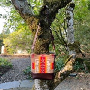 May include: A tan and brown shoulder bag with a geometric pattern in red, orange, and white. The bag has a brown leather bottom and a brown leather strap. The bag is hanging from a tree branch.