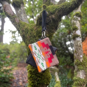 May include: A small, square clutch with a black base and a patterned top featuring geometric designs in red, orange, green, and white. The clutch is suspended from a black wrist strap attached to a tree branch.
