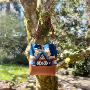 May include: A blue and brown shoulder bag with a geometric pattern and a brown leather bottom. The bag has a brown leather strap and is hanging from a tree branch. The bag's design includes white, tan, and orange accents.