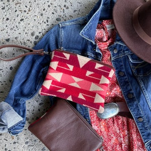 May include: A red and pink patterned clutch with a brown leather clutch with a strap. The clutches are on top of a red floral dress and a denim jacket.