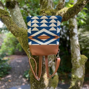 May include: A small blue and white patterned backpack with brown leather straps and a brown leather bottom. The backpack is hanging from a tree branch.