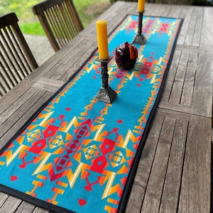 May include: A turquoise table runner with a Southwestern geometric pattern in red, orange, and yellow. The runner is bordered with black trim and is displayed on a wooden table. Two ornate candle holders with yellow candles and a decorative jar are on the runner.
