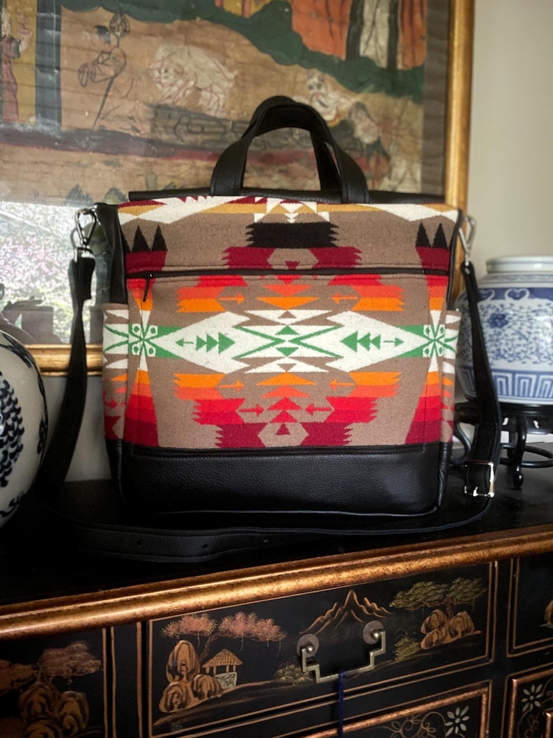 Puede incluir: Un bolso tote de patr&oacute;n marr&oacute;n y negro con fondo de cuero negro y una correa larga ajustable. El bolso tiene un bolsillo con cremallera en la parte delantera.