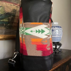 Puede incluir: Una mochila de cuero negro con un patr&oacute;n tejido de colores. La mochila tiene un asa superior y un cierre de cremallera.