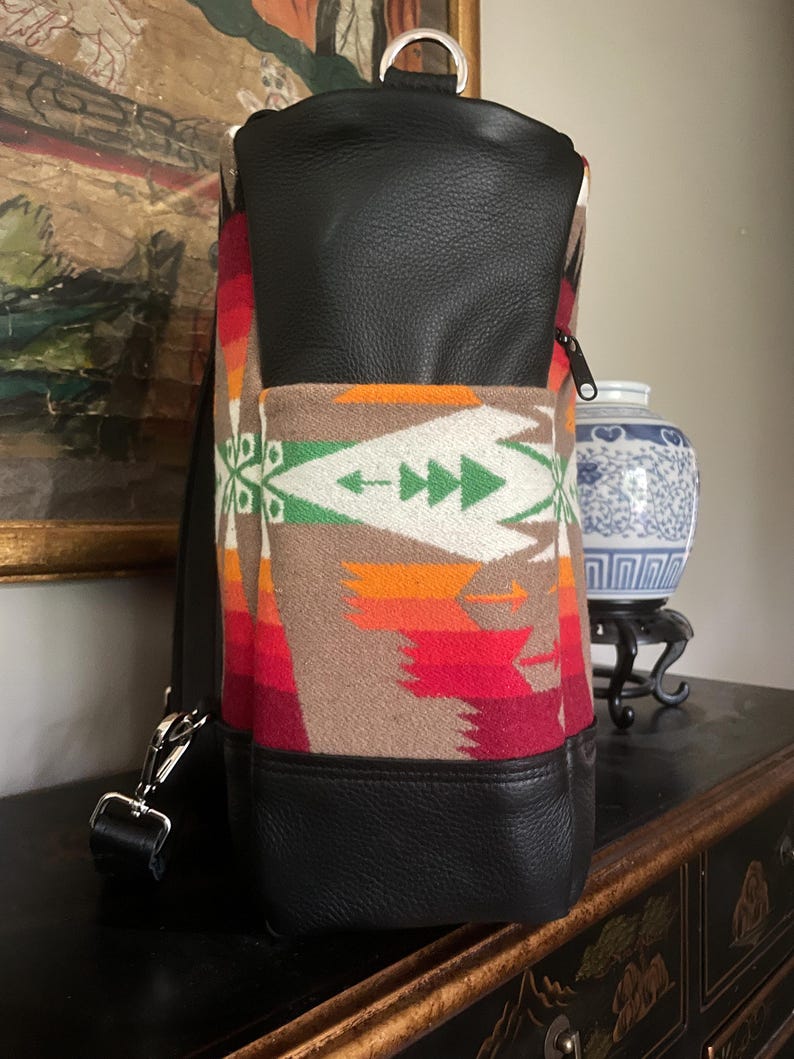 Puede incluir: Una mochila de cuero negro con un patr&oacute;n tejido de colores. La mochila tiene un asa superior y un cierre de cremallera.