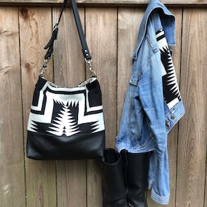May include: A black leather tote bag with a white and black geometric pattern. The bag has a long strap and is hanging on a wooden fence. There are also two black boots hanging on the fence.
