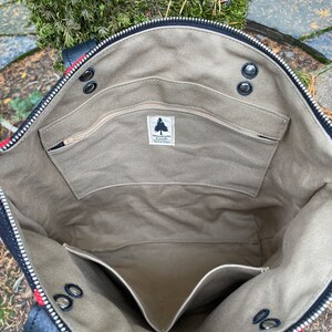 K&ouml;nnte beinhalten: Das Innere einer schwarzen Canvas-Tragetasche mit Rei&szlig;verschluss. Die Tasche hat ein gro&szlig;es Hauptfach und eine kleinere Rei&szlig;verschlusstasche. Die Tasche ist mit einem beigefarbenen Stoff gef&uuml;ttert und tr&auml;gt ein Etikett mit der Aufschrift "Woodside Goods".