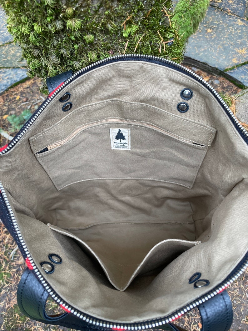 K&ouml;nnte beinhalten: Das Innere einer schwarzen Canvas-Tragetasche mit Rei&szlig;verschluss. Die Tasche hat ein gro&szlig;es Hauptfach und eine kleinere Rei&szlig;verschlusstasche. Die Tasche ist mit einem beigefarbenen Stoff gef&uuml;ttert und tr&auml;gt ein Etikett mit der Aufschrift "Woodside Goods".