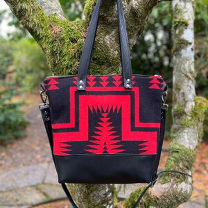 K&ouml;nnte beinhalten: Eine schwarze und rote Tragetasche mit geometrischem Muster. Die Tasche hat einen schwarzen Lederboden und einen langen schwarzen Riemen.