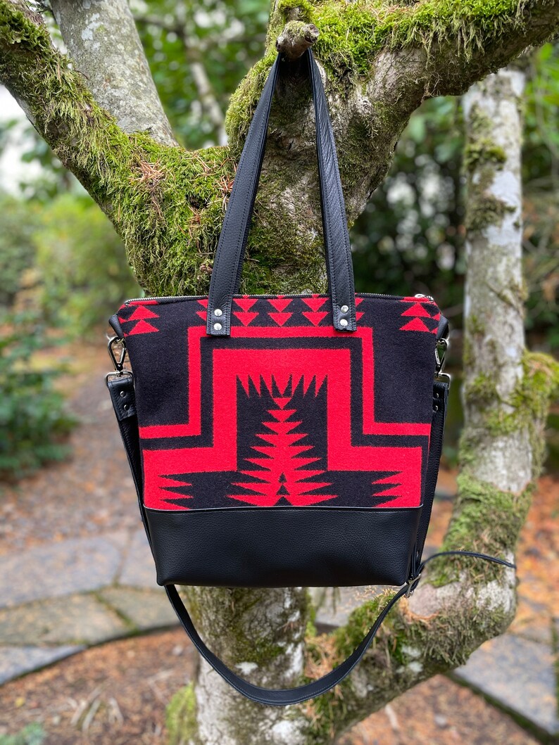 K&ouml;nnte beinhalten: Eine schwarze und rote Tragetasche mit geometrischem Muster. Die Tasche hat einen schwarzen Lederboden und einen langen schwarzen Riemen.