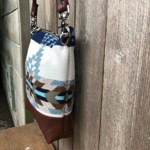 May include: A white and blue patterned crossbody bag with a brown leather bottom and strap. The bag has a silver clasp and a zipper closure.