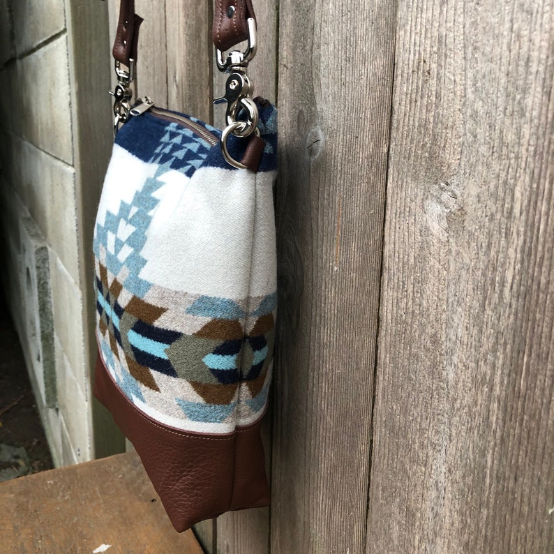 May include: A white and blue patterned crossbody bag with a brown leather bottom and strap. The bag has a silver clasp and a zipper closure.