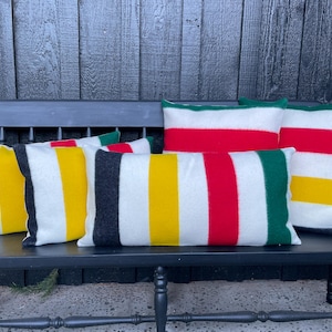 May include: A collection of rectangular throw pillows with bold, colorful stripes. The pillows feature stripes of red, yellow, green, and black against a white background. They are arranged on a black bench, creating a vibrant and cozy aesthetic.