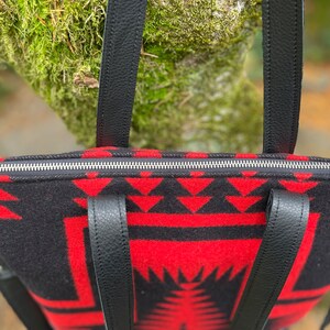 K&ouml;nnte beinhalten: Eine schwarze und rote gemusterte Tragetasche mit schwarzen Lederriemen und einem silbernen Rei&szlig;verschluss. Die Tasche hat ein geometrisches Design.