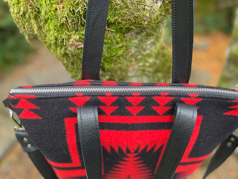 K&ouml;nnte beinhalten: Eine schwarze und rote gemusterte Tragetasche mit schwarzen Lederriemen und einem silbernen Rei&szlig;verschluss. Die Tasche hat ein geometrisches Design.