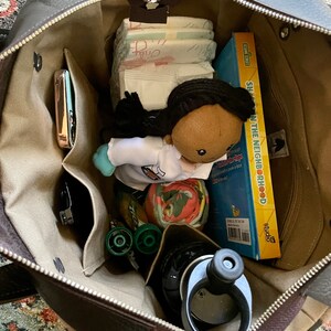 Puede incluir: Una bolsa de mano de cuero marr&oacute;n abierta y llena de varios art&iacute;culos, incluyendo una mu&ntilde;eca, un libro titulado "Sesame Street:  Show in the Neighborhood", una botella de agua, pa&ntilde;ales y un tel&eacute;fono en un bolsillo.